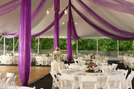 Purple sashes hang from the ceiling of a white tent set up for a backyard wedding.